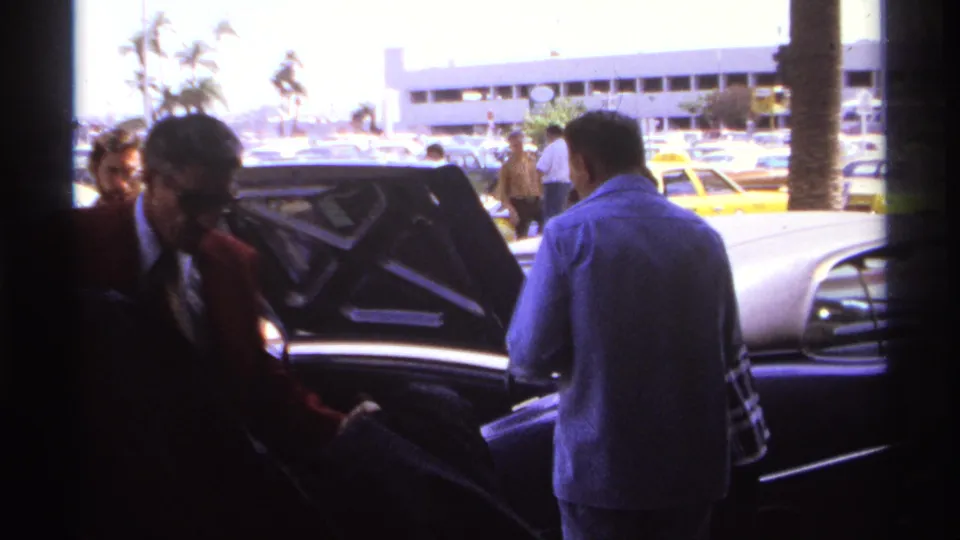 Home-movie frame of men beside an open sedan trunk at a sunny 1974 airport curb in La Paz, Mexico.