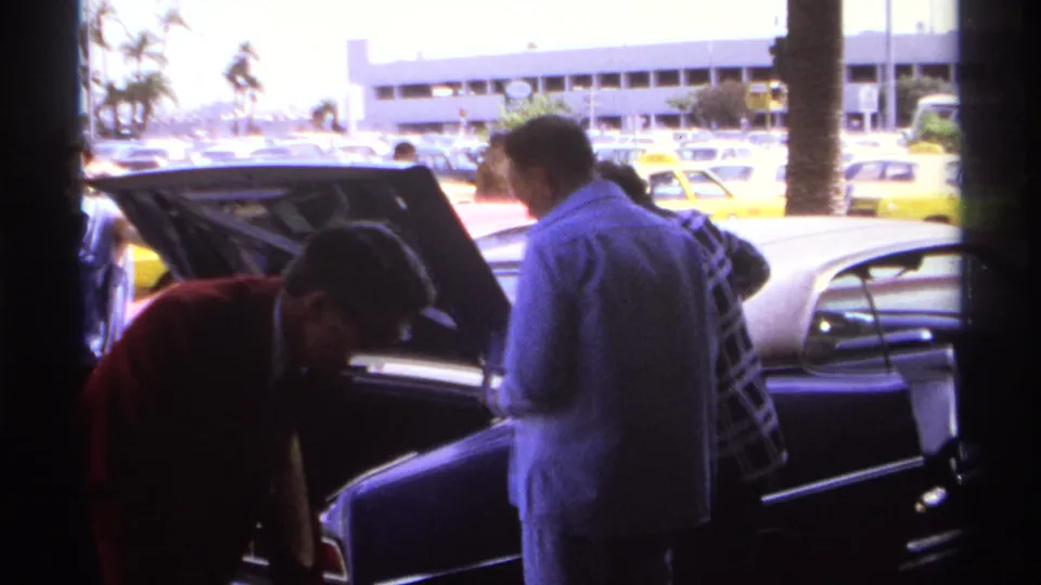 At the La Paz airport curb, a man in a red jacket bends near an open trunk as two men stand beside the sedan.