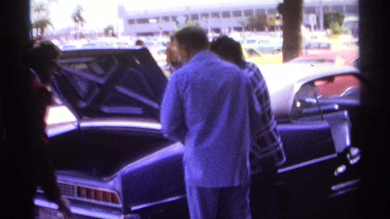 Travelers stand near a dark car with its trunk open outside an airport terminal.