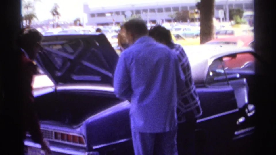 1974 La Paz, Mexico airport scene with people near a dark sedan, open trunk, parked cars, and bright exterior light.
