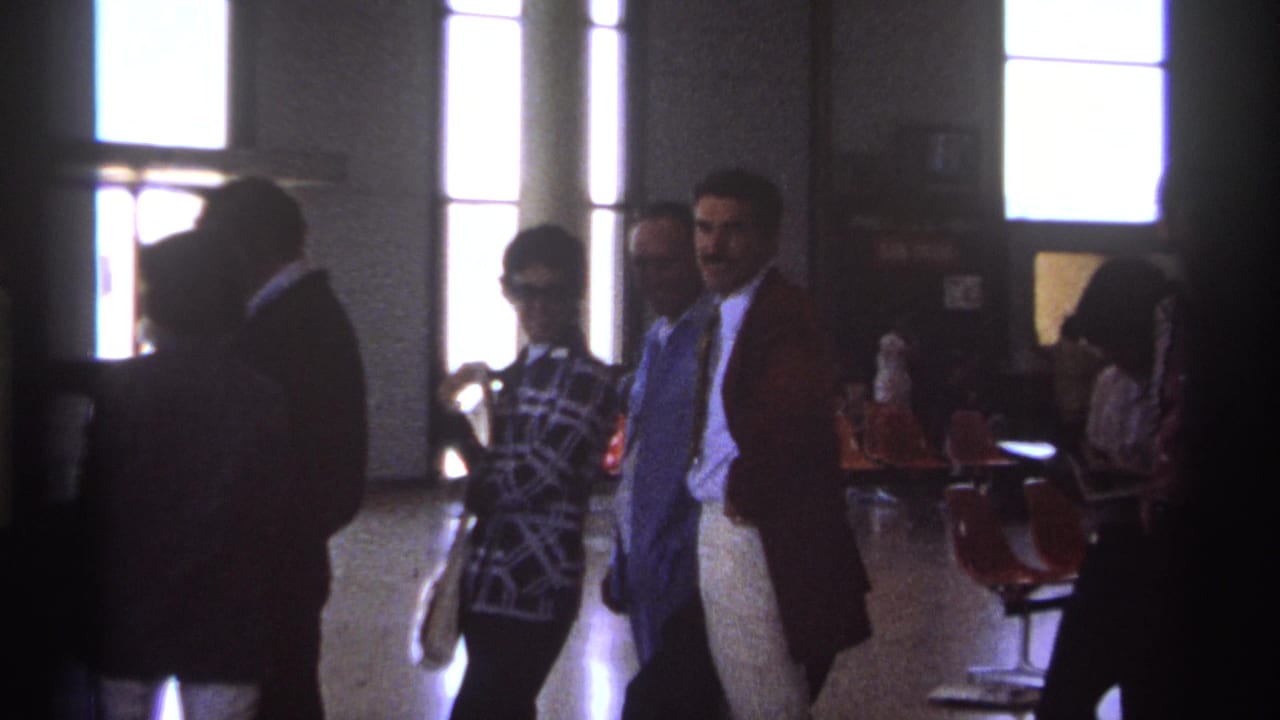 Passengers walk and wait inside a dim airport terminal with bright windows behind them.