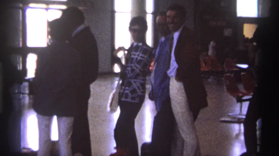 1974 La Paz terminal view with a woman in sunglasses, men in suits, red seating, and bright window light.