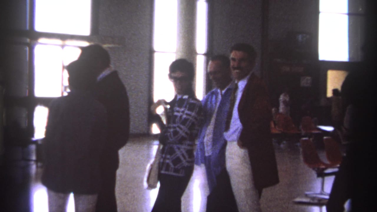 Adults stand and talk in a public interior beside windows and orange seating.