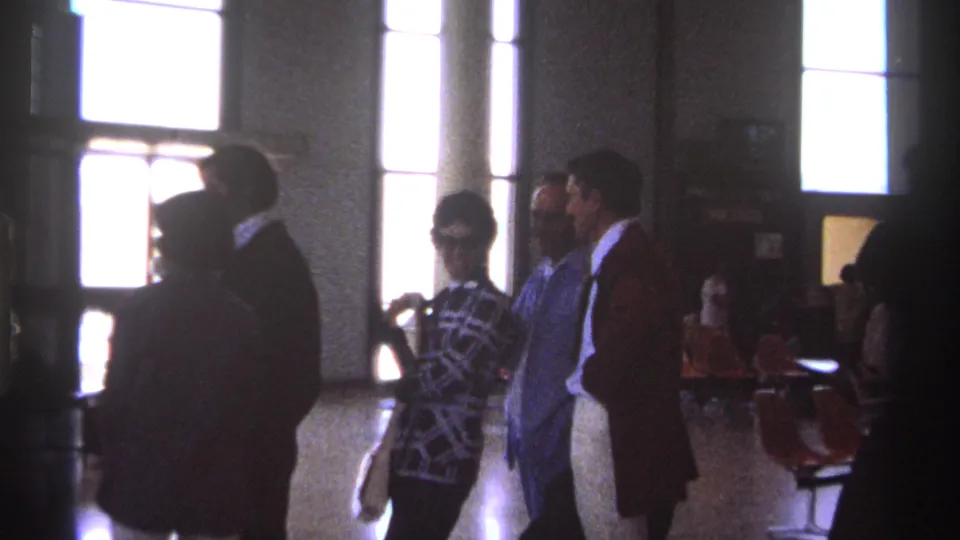 1974 La Paz, Mexico home-movie still showing a small queue in a public interior, with period clothing, columns, and orange waiting chairs.