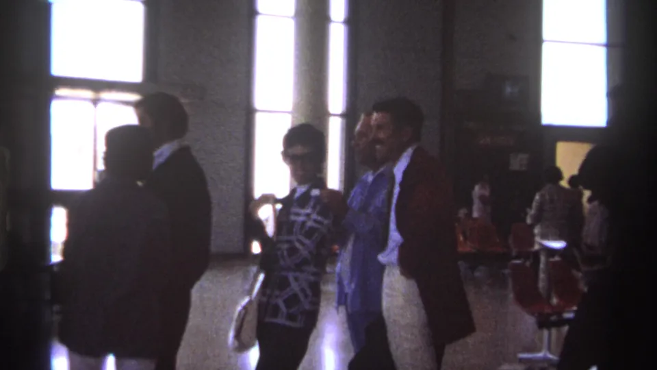 1974 La Paz, Mexico frame showing a smiling man in a suit among adults waiting in a public hall with orange chairs and strong window light.