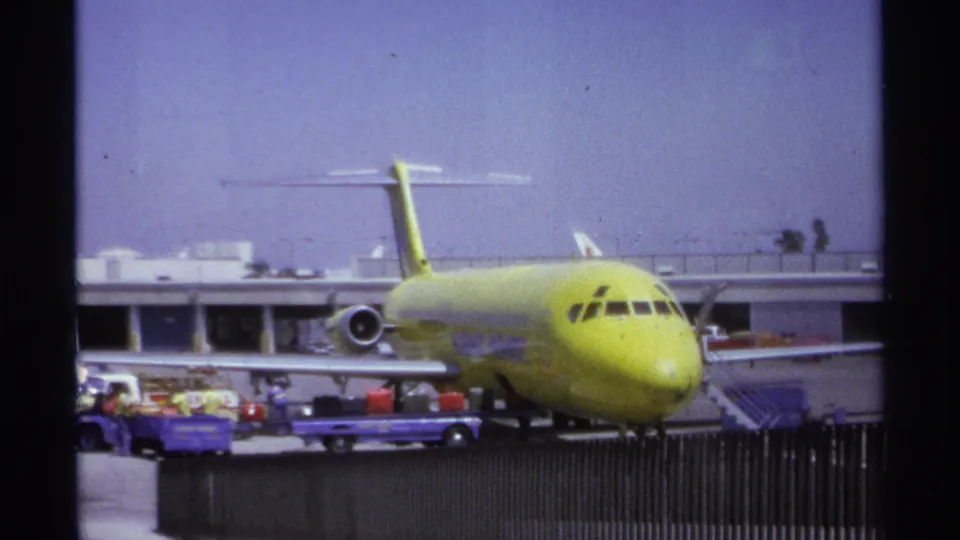 1974 La Paz airport view of a yellow plane with baggage carts, workers, fencing, and terminal structures visible around it.
