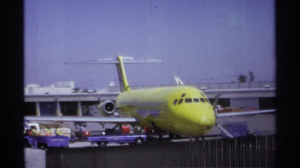 A lime-green commercial airplane sits at a La Paz airport gate in 1974 as distant workers handle baggage beside the aircraft.