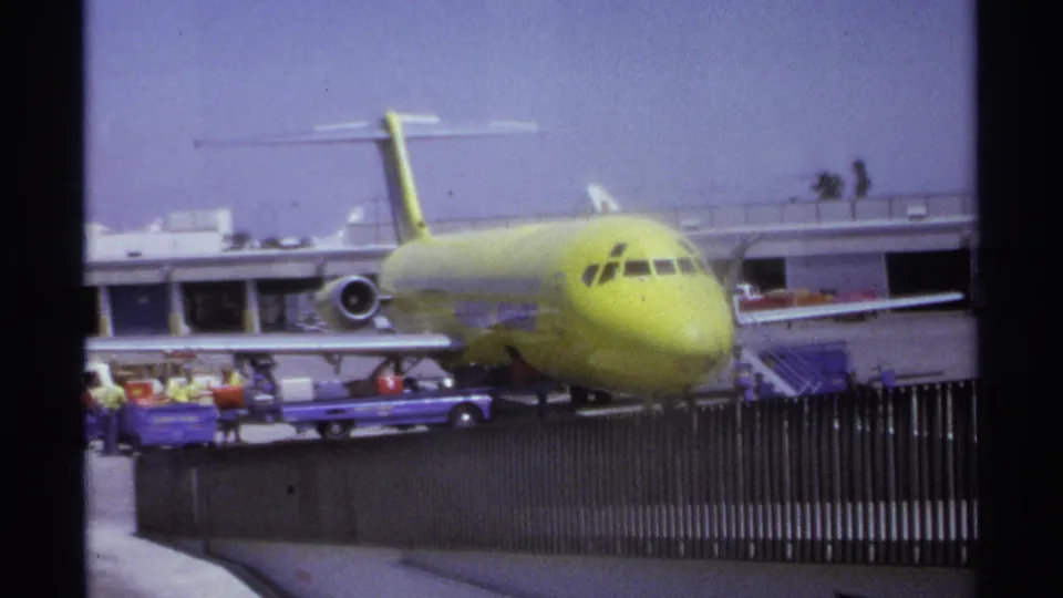 1974 home-movie view in La Paz showing the lime-green airplane from slightly closer framing with luggage carts and terminal behind it.