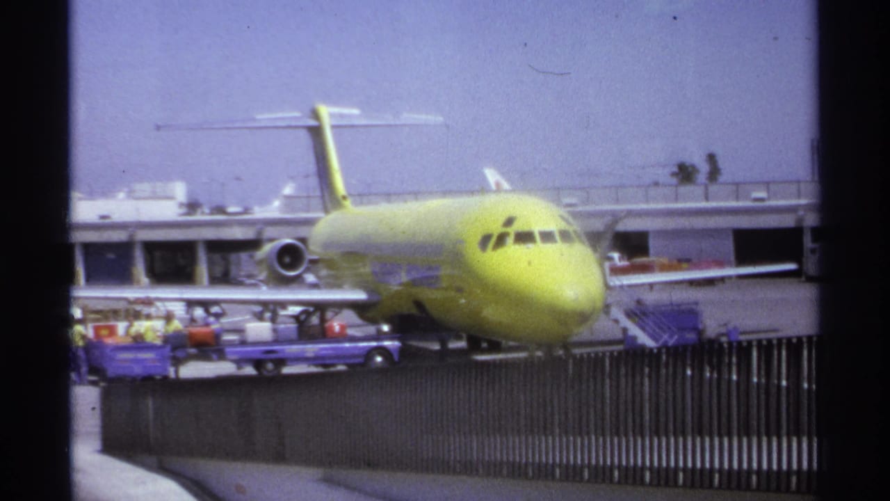 Yellow jet parked at an airport gate with cargo carts and ground crew behind a fence.