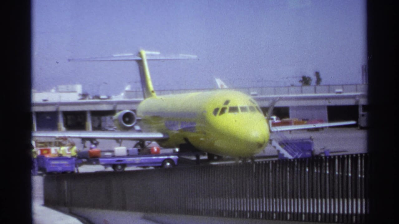 Yellow airplane beside the terminal with cargo carts, stairs, and workers in the foreground.
