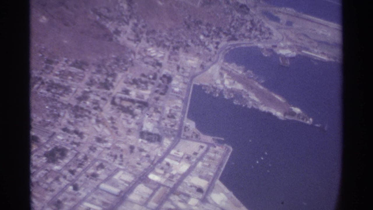 Aerial view of a coastal city, harbor water, shoreline roads, and a pier seen through aircraft-window framing.