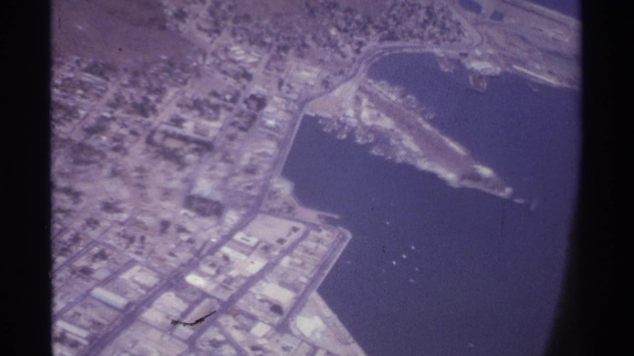 Aerial harbor view with shoreline buildings, a curved road, pier structures, and open water below.