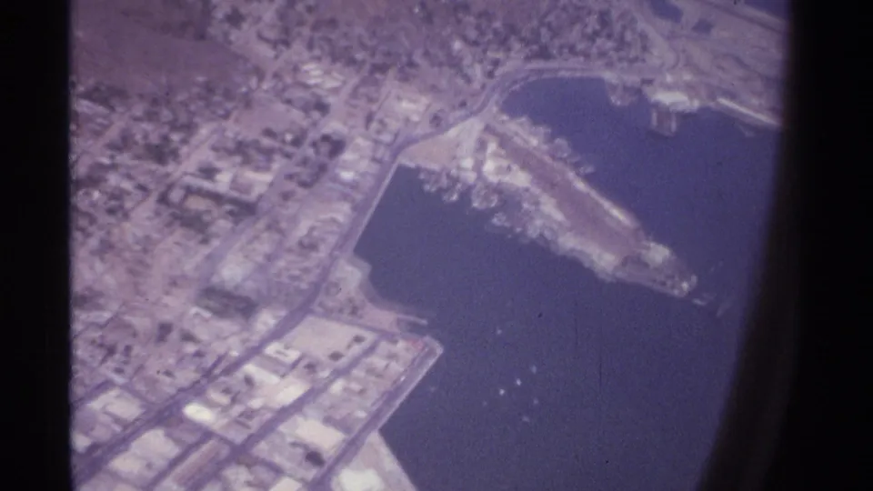 A 1974 8mm aerial view over La Paz, Mexico, with waterfront roads, harbor structures, urban blocks, and small boats or light points in the bay.