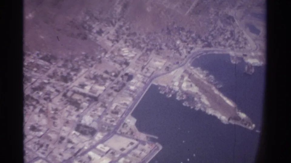 1974 aircraft-window view of La Paz, Mexico, showing the harbor basin, shoreline streets, low-rise city fabric, and several small boats on the water.