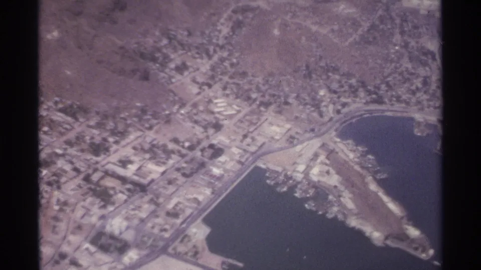 Aerial 8mm frame from La Paz, Mexico, with a harbor basin, shoreline road, clustered buildings, and dry terrain inland.