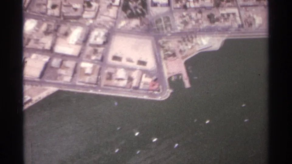 1974 La Paz aerial frame showing waterfront buildings, a harbor edge, pier structures, and small boats in dark water.
