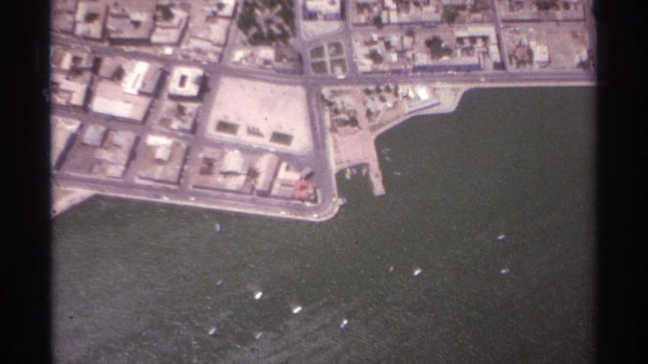 Aerial view of a waterfront city grid beside dark harbor water with small boats offshore.