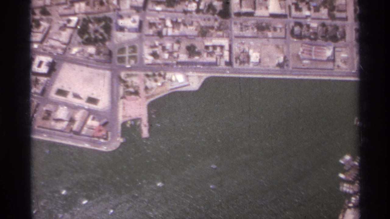 Aerial view of coastal blocks, streets, harbor water, and a marina or dock area at frame edge.