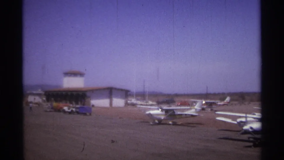 1974 La Paz, Mexico airfield frame showing parked light planes, open tarmac, vehicles, and a control tower in soft 8mm color.