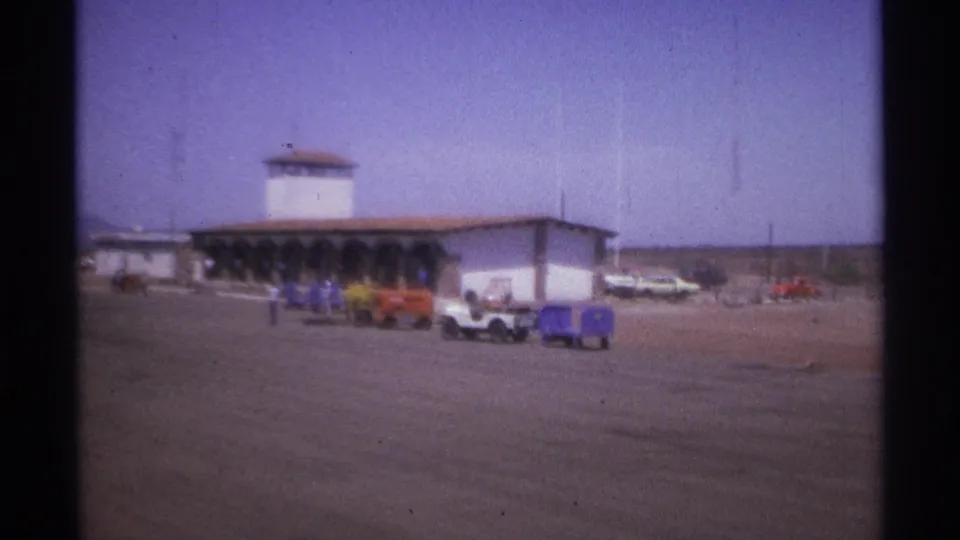 1974 home-movie view of a La Paz airport terminal with distant people, classic cars, blue luggage carts, and open pavement.