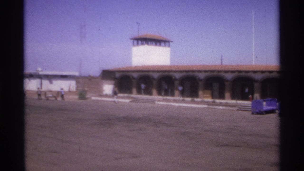 Building with arched arcade and white tower, with people walking and a blue cart at right.