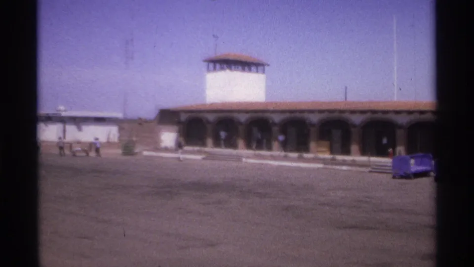 1974 La Paz, Mexico frame of a sunlit arched facade, white tower, pedestrians, and a blue luggage cart.