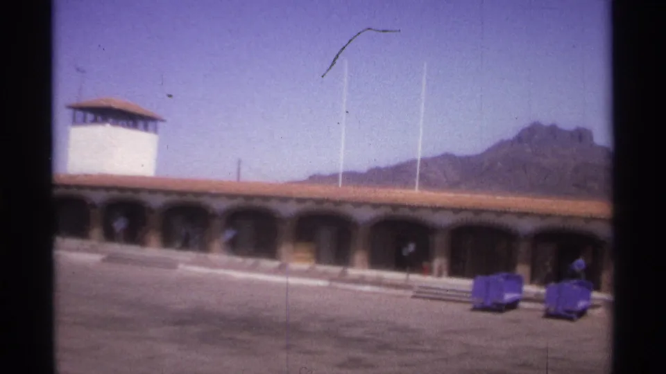 1974 La Paz, Mexico 8mm frame showing an arched building, white tower, open paved plaza, luggage carts, and distant mountains.