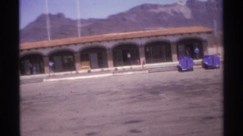 1974 La Paz, Mexico 8mm frame looking toward an arched colonnade with purple luggage carts, steps, flagpoles, and mountains behind.