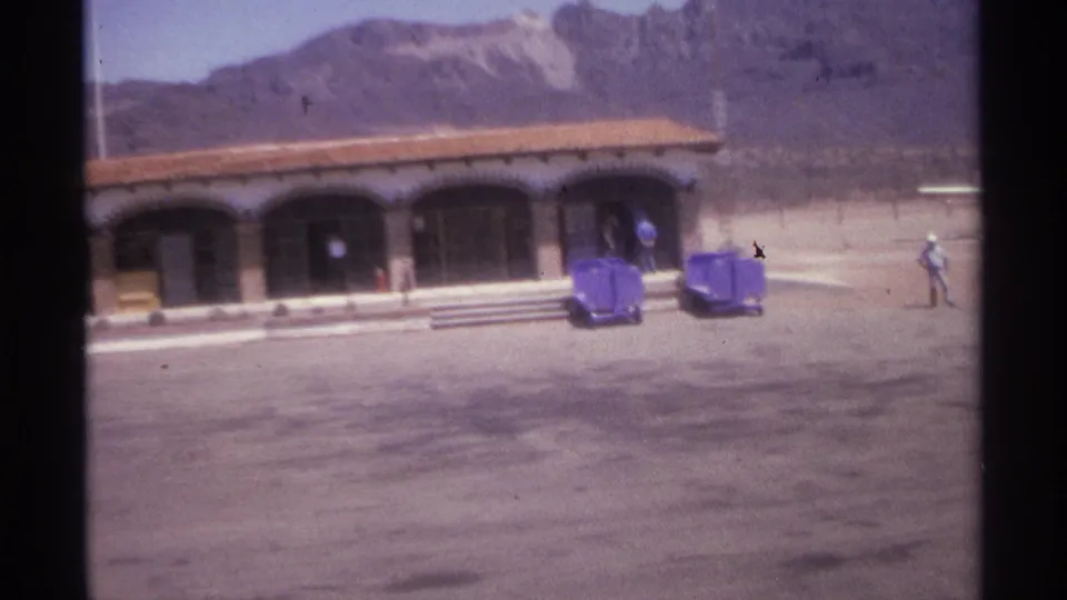 1974 La Paz, Mexico frame showing an arched building, purple carts, broad pavement, mountains, and a pedestrian crossing at right.