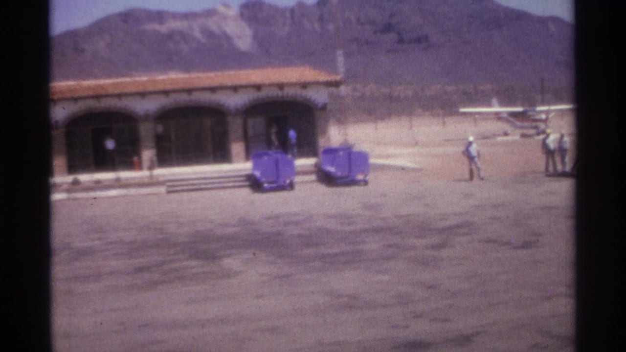 1974 La Paz, Mexico 8mm frame showing an arched desert building, mountain backdrop, people, carts, and a small aircraft beyond the forecourt.