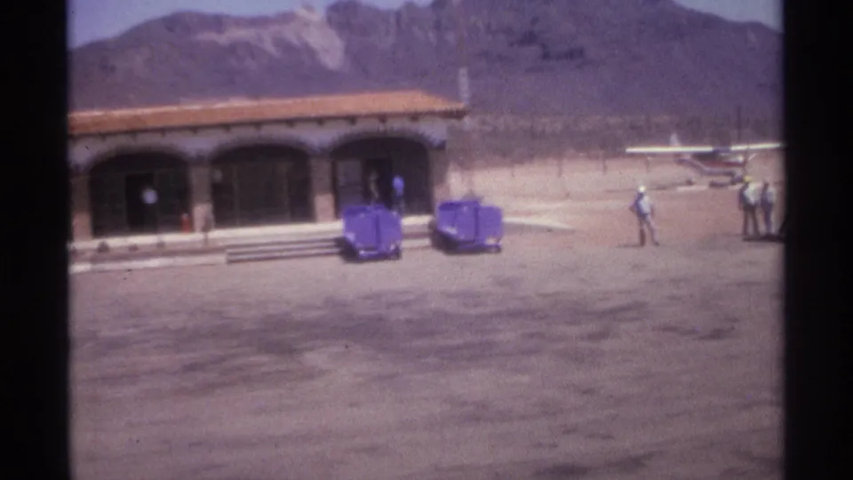 1974 La Paz, Mexico 8mm frame showing an arched desert building, mountain backdrop, people, carts, and a small aircraft beyond the forecourt.