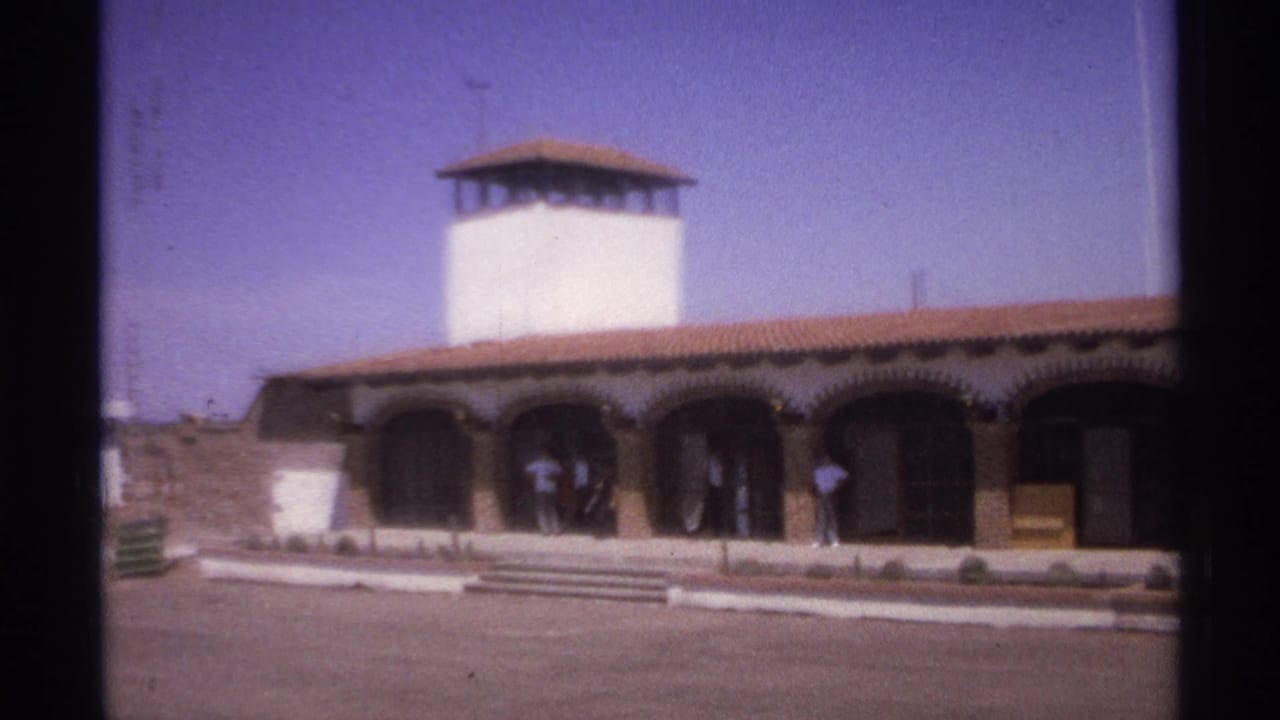 1974 La Paz, Mexico frame of a low arched facade with a white tower, terracotta roof, paved forecourt, and adults by the entrance.