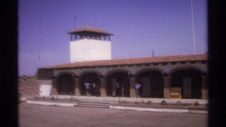 1974 La Paz, Mexico 8mm view of a white tower above an arched public-looking building, with adults near the shaded entryway.
