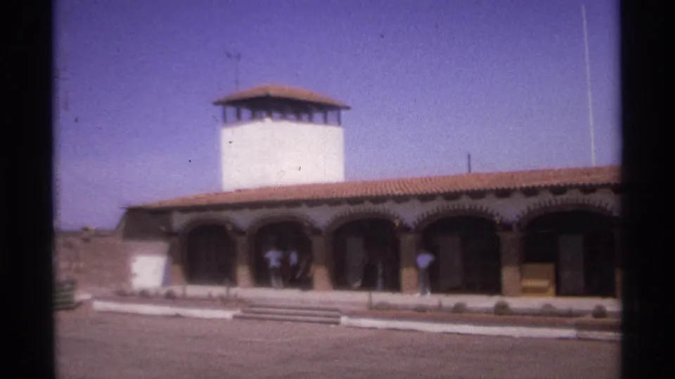 1974 La Paz, Mexico frame showing the arched exterior, white tower, tiled roof, paved forecourt, and adults by the shaded doorway.