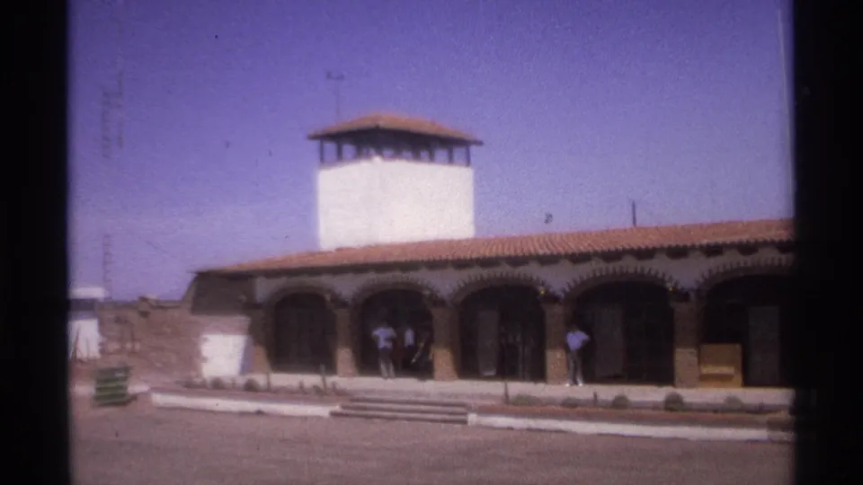 1974 La Paz home-movie frame showing a white arched building with a tower, red tile roof, paved foreground, and people near the doors.