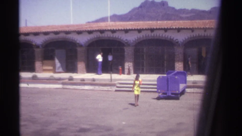 1974 La Paz frame with a woman in a yellow dress crossing the courtyard outside the arched building, with a purple cart nearby.