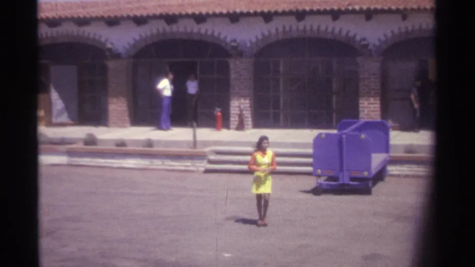 1974 La Paz home-movie frame showing the woman closer to camera in a yellow dress, with arched doors, steps, and people behind her.