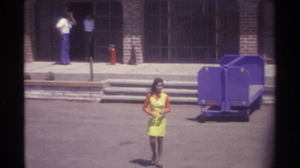 1974 La Paz frame showing a closer courtyard view of the woman in yellow, with arched doors, steps, people, and the purple cart in back.