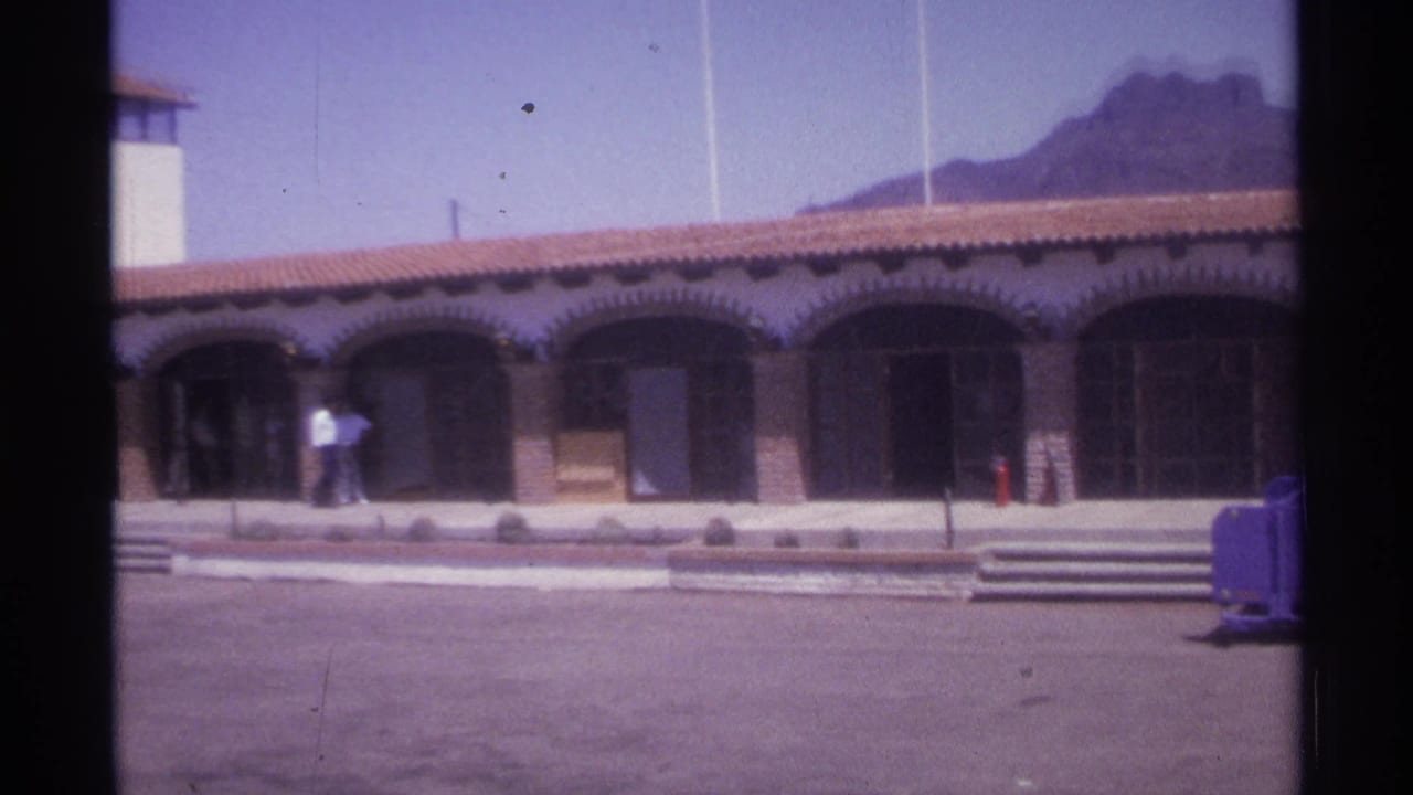 Arched building with red tile roof, pedestrians, tower, and mountain in the background.