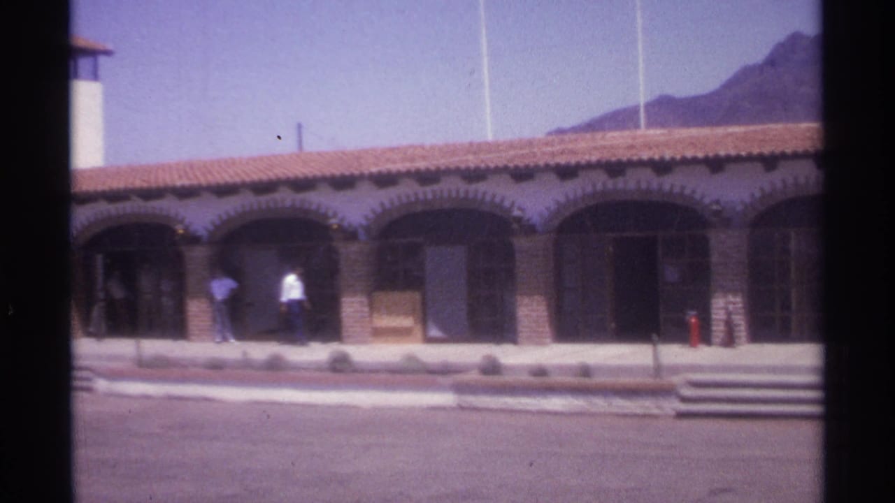 1974 La Paz frame with pedestrians moving along an arched walkway beneath a red tile roof, with mountains behind.