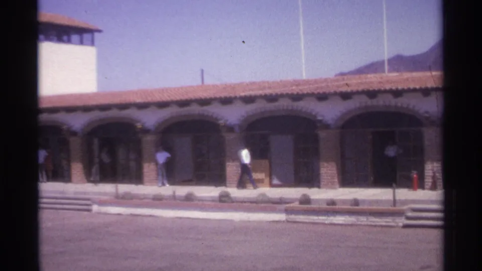 1974 La Paz 8mm frame showing the tower end of an arched building as people stand and walk near the storefronts.