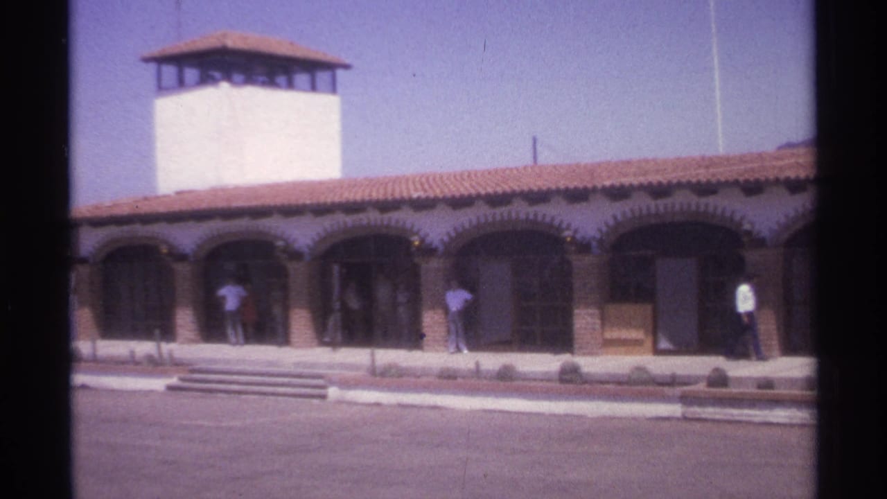 Wide view of a Spanish-style building with a white tower, arched entries, and people standing near the doors.