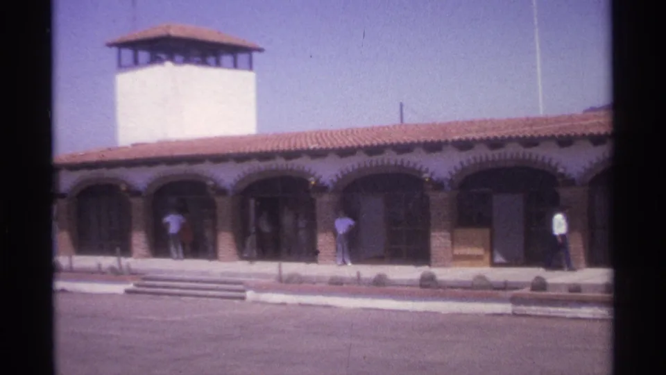 1974 La Paz, Mexico 8mm frame showing a red-tiled building exterior with arched entrances, a white tower, front steps, and several people outside.