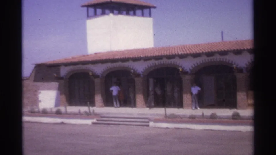 Closer 8mm frame of the 1974 La Paz exterior, with arched bays, a red-tiled roof, white tower, steps, and people by the doors.