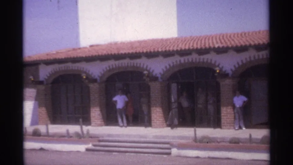 Tight 1974 La Paz home-movie frame showing people outside the shaded arched entrances beneath a red-tiled roof and white tower.