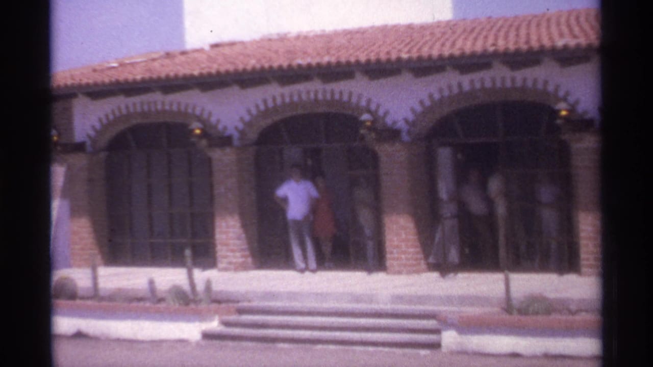 Several adults stand near arched doors on a sunlit Spanish-style building.