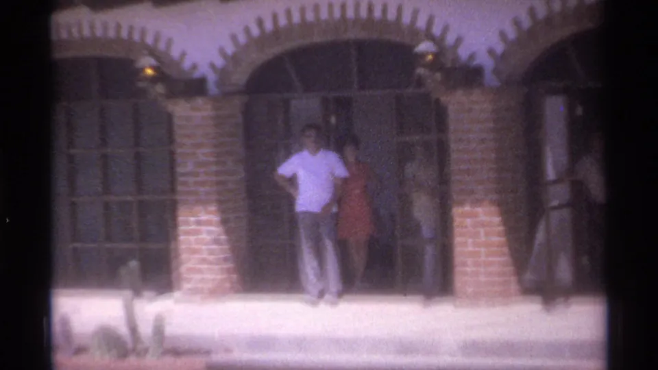 1974 La Paz frame with a man in a light shirt and a woman in a red dress standing at a shaded arched entrance.