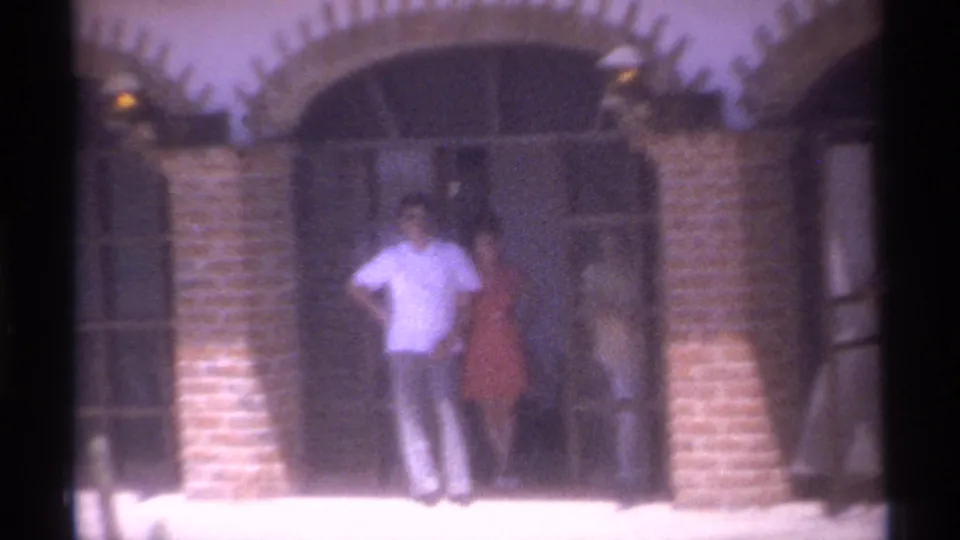 1974 La Paz home-movie frame centered on adults standing before a dark arched doorway with brick columns and metal doors.
