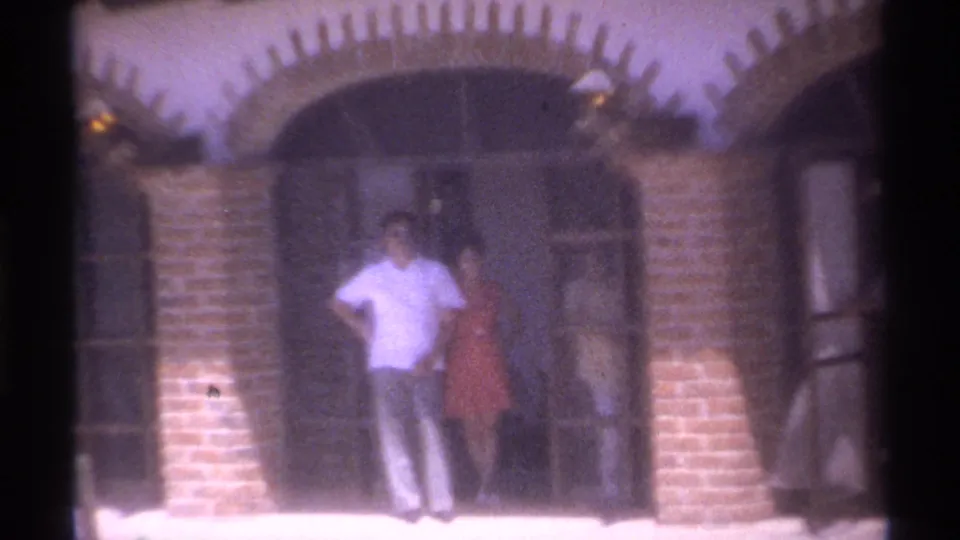 Final 1974 La Paz frame showing adults at a shaded arched entrance, with brick columns, metal doors, and warm 8mm color.