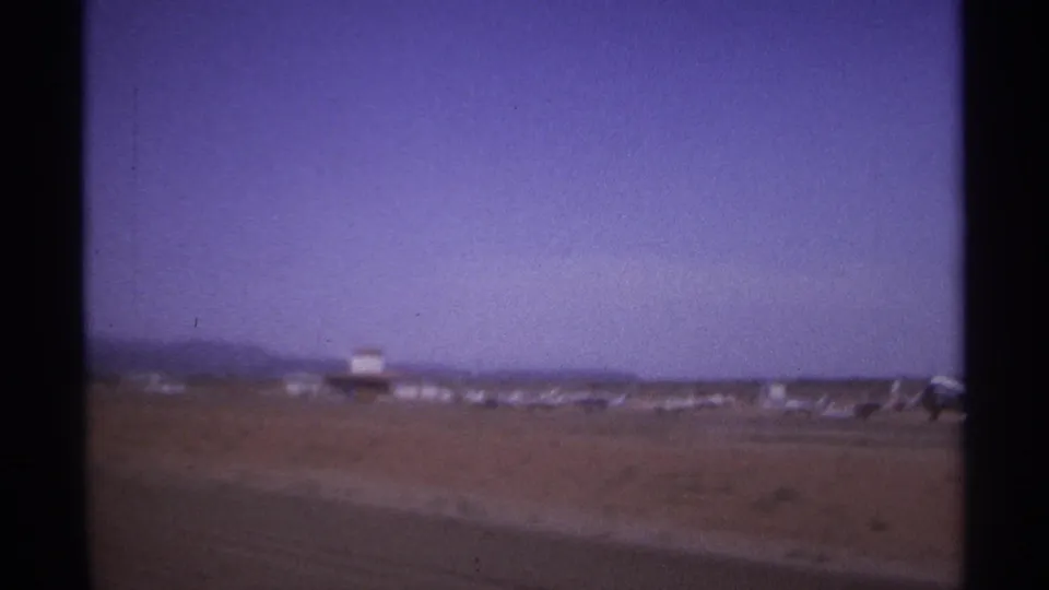 The frame holds on the desert airfield, with distant equipment and a partial aircraft shape at the right edge.